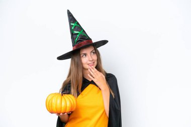 Beyaz arka planda izole edilmiş bir balkabağı tutan cadı kostümü giymiş genç beyaz kadın gülümserken yukarı bakıyor.