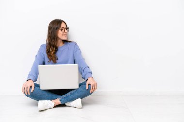 Young caucasian woman with a laptop sitting on the floor isolated on white background looking side