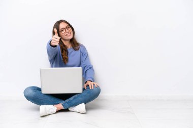 Young caucasian woman with a laptop sitting on the floor isolated on white background with thumbs up because something good has happened