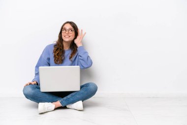 Dizüstü bilgisayarı olan genç beyaz bir kadın, beyaz arka planda izole bir şekilde oturuyor ve elini kulağına koyarak bir şeyler dinliyor.