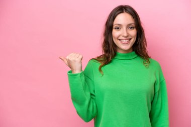 Pembe arka planda izole edilmiş genç beyaz kadın bir ürünü sunmak için kenarı işaret ediyor.