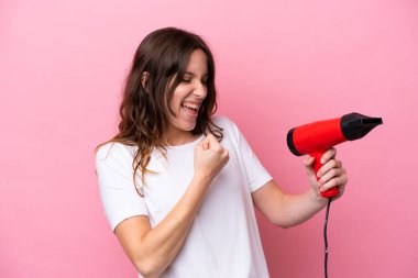 Pembe arka planda saç kurutma makinesi tutan beyaz bir kadın zaferi kutluyor.