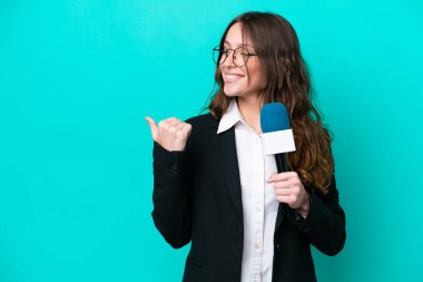 Mavi arka planda izole edilmiş genç TV sunucusu kadın bir ürünü sunmak için kenarı işaret ediyor