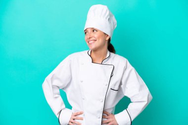 Kafkasyalı genç aşçı kadın mavi arka planda izole edilmiş, kolları kalçasında ve gülümsüyor.