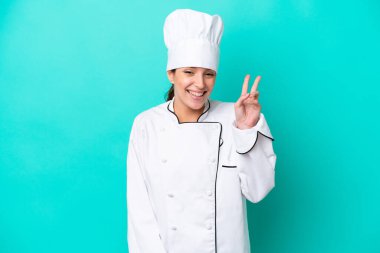 Kafkasyalı genç aşçı kadın mavi arka planda izole edilmiş gülümsüyor ve zafer işareti gösteriyor.