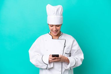 Kafkasyalı genç aşçı kadın mavi arka planda izole edilmiş cep telefonuyla bir mesaj gönderiyor.
