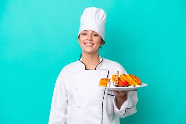 Kafkasyalı genç aşçı kadın, elinde waffle tutarken mavi arka planda izole bir şekilde gülümsüyor.
