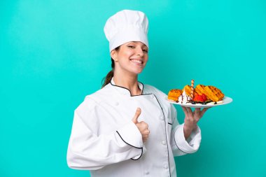 Kafkasyalı genç aşçı kadın, mavi arka planda gözleme tutuyor. Başparmakları havada, çünkü iyi bir şey oldu.