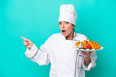Kafkasyalı genç aşçı kadın elinde waffle tutuyor mavi arka planda izole edilmiş şaşırmış ve işaret ediyor.
