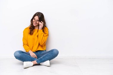 Beyaz duvarda, gözlüklü ve mutlu bir şekilde yerde oturan beyaz bir kadın.