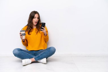 Beyaz duvarda izole bir şekilde yerde oturan beyaz bir kadın elinde kahve ve bir cep telefonu tutuyor.