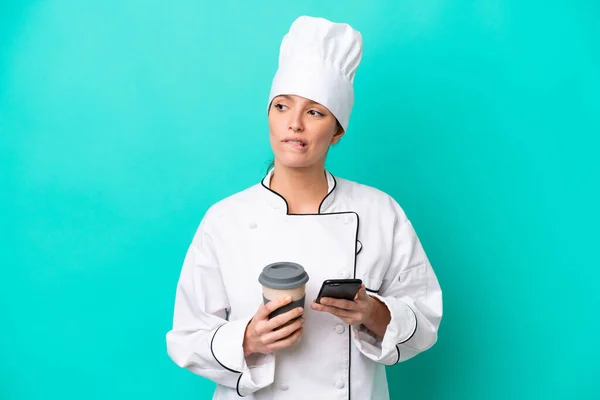 Young caucasian chef woman isolated on blue background holding coffee to take away and a mobile while thinking something