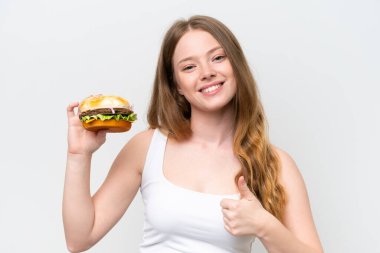 Genç ve güzel bir kadın beyaz arka planda izole edilmiş bir burger tutuyor. Başparmakları havada çünkü iyi bir şey oldu.