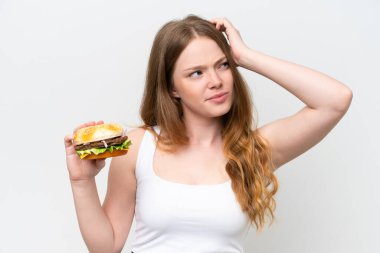 Beyaz arka planda izole edilmiş bir burger tutan genç ve güzel bir kadın. Şüpheleri var ve kafası karışmış bir yüz ifadesi var.