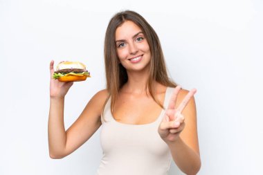 Beyaz arka planda izole edilmiş bir burger tutan genç ve güzel bir kadın gülümsüyor ve zafer işareti gösteriyor.