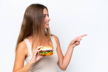 Beyaz arka planda izole edilmiş bir burger tutan genç ve güzel bir kadın bir ürünü sunmak için kenarı işaret ediyor.