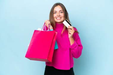 Mavi arka planda izole edilmiş genç bir kadın elinde alışveriş çantaları ve bir kredi kartı tutuyor.