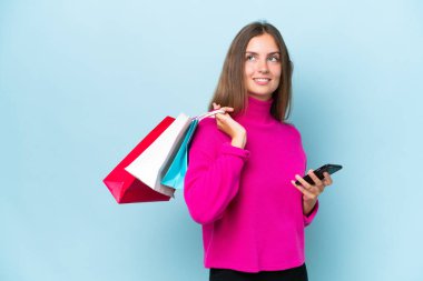 Mavi arka planda izole edilmiş genç bir kadın elinde alışveriş çantaları ve cep telefonu tutuyor.