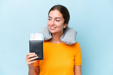 Şişirilebilir seyahat yastığı olan genç ve güzel bir kadın mavi arka planda izole edilmiş, yana bakıyor ve gülümsüyor.