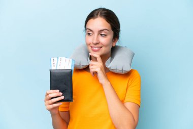 Şişirilebilir seyahat yastığı olan genç ve güzel bir kadın mavi arka planda izole edilmiş, yana bakıyor ve gülümsüyor.