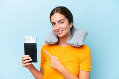 Şişirilebilir seyahat yastığı olan genç ve güzel bir kadın mavi arka planda izole edilmiş bir şekilde bir ürün sunmak için kenarı işaret ediyor.