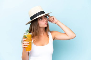 Genç ve güzel bir kadın mavi arka planda izole edilmiş bir kokteyl tutuyor. Şüpheleri var ve kafası karışmış bir yüz ifadesi var.