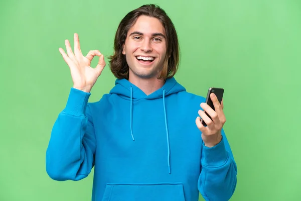 携帯電話を使用して緑のクロマの背景に隔離された若いハンサムな男とOkサインをします — ストック写真