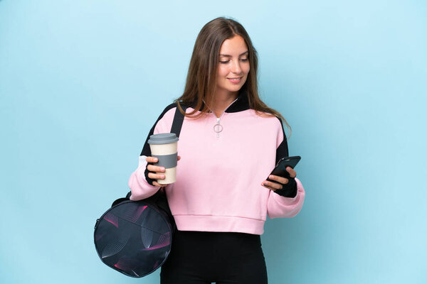 Young sport woman with sport bag isolated on blue background holding coffee to take away and a mobile