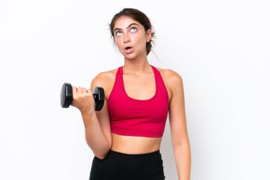 Beyaz arka planda halter kaldıran genç beyaz kadın şaşırmış bir ifadeyle yukarı bakıyor.