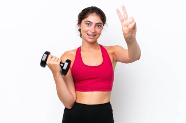 Beyaz arka planda halter kaldıran genç beyaz kadın gülümsüyor ve zafer işareti gösteriyor.