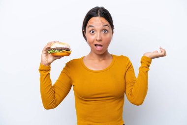 Beyaz arka planda yüzü şoke olmuş bir hamburger tutan genç İspanyol kadın.