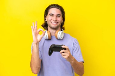Sarı arka planda izole edilmiş bir bilgisayar oyunu kontrolörüyle oynayan yakışıklı beyaz bir adam parmaklarıyla tamam işareti gösteriyor.