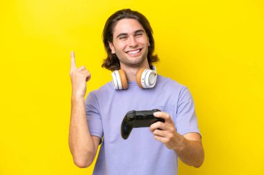Sarı arka planda izole edilmiş bir bilgisayar oyunu kontrolörüyle oynayan yakışıklı beyaz bir adam en iyinin işareti olarak parmağını kaldırıyor.