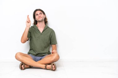 Genç yakışıklı adam beyaz arka planda izole bir şekilde yerde oturuyor. Parmaklarını çapraz yapıp en iyisini diliyor.
