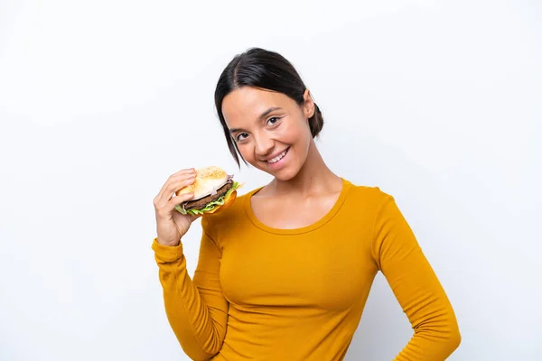 Młoda Latynoska Trzymająca Hamburgera Białym Tle Pozująca Rękami Biodrze Uśmiechnięta — Zdjęcie stockowe