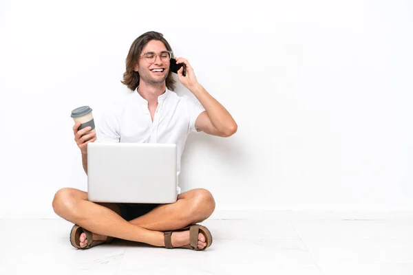 Young handsome man with a laptop sitting on the floor isolated on white background holding coffee to take away and a mobile