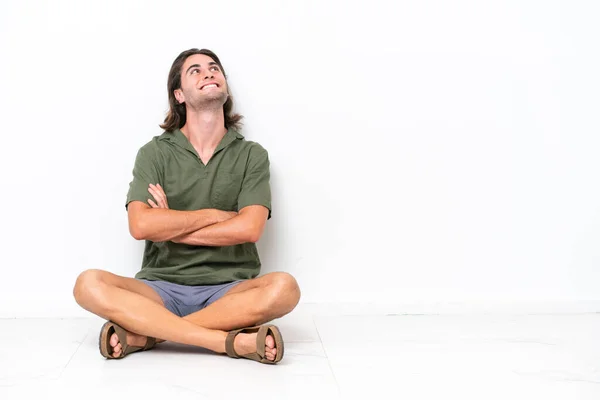 Mladý Pohledný Muž Sedí Podlaze Izolované Bílém Pozadí Vzhlíží Usmívá — Stock fotografie