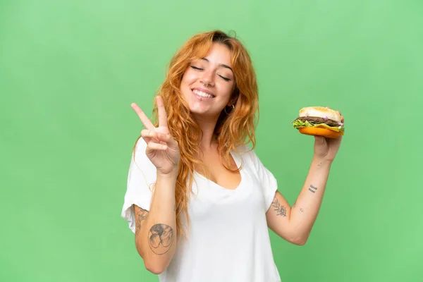 緑の画面に隔離されたハンバーガーを保持若い白人女性クロマキー背景笑顔と勝利の兆候を示す — ストック写真