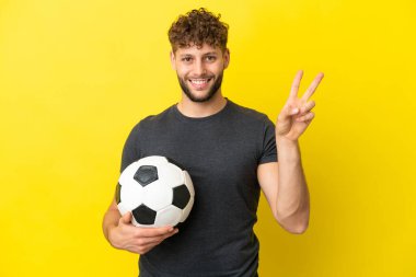 Yakışıklı genç futbolcu sarı arka planda izole edilmiş gülümsüyor ve zafer işareti gösteriyor.