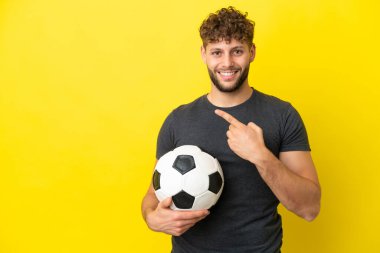 Yakışıklı genç futbolcu sarı arka planda izole edilmiş bir şekilde bir ürün sunmak için kenarı işaret ediyor.