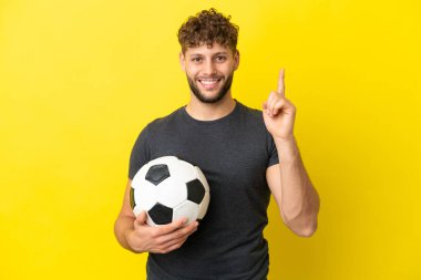 Yakışıklı genç futbolcu sarı arka planda izole edilmiş ve en iyinin işareti olarak parmağını kaldırıyor.