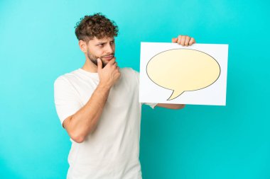 Genç beyaz adam mavi arka planda izole edilmiş. Elinde konuşma balonu ikonu olan bir levha var ve düşünüyor.