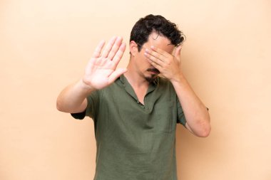 Beyazlı genç bir adam bej arka planda izole edilmiş. Durdurma hareketi yapıyor ve yüzünü kapatıyor.