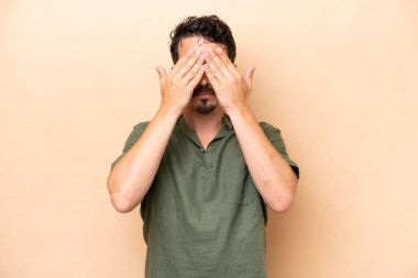 Beyazlı genç bir adam bej arka planda izole edilmiş, gözleri elle kapatılmış.