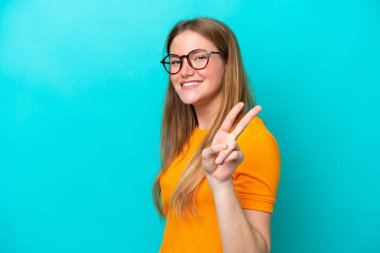 Mavi arka planda izole edilmiş genç beyaz kadın gülümsüyor ve zafer işareti gösteriyor.