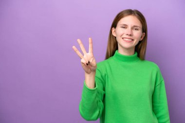 Mor arka planda izole edilmiş genç İngiliz kadın mutlu ve parmaklarıyla üçe kadar sayıyor.