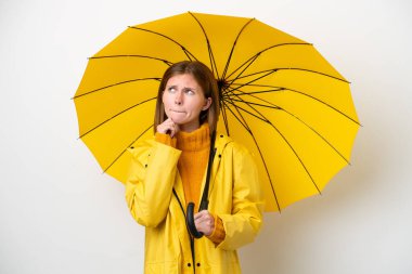 Young English woman with rainproof coat and umbrella isolated on white background having doubts and thinking