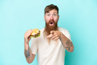 Kızıl saçlı uzun sakallı bir adam elinde bir burger tutuyor mavi arka planda izole edilmiş şaşırmış ve ön tarafı işaret ediyor