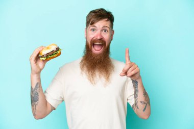 Kızıl saçlı uzun sakallı bir adam elinde mavi arka planda izole edilmiş bir burger tutuyor. Harika bir fikir.