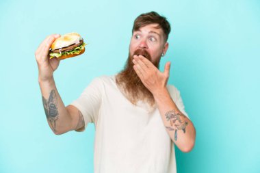 Uzun sakallı kızıl saçlı adam mavi arka planda bir burger tutuyor. Şaşırmış ve şok olmuş bir yüz ifadesi var.